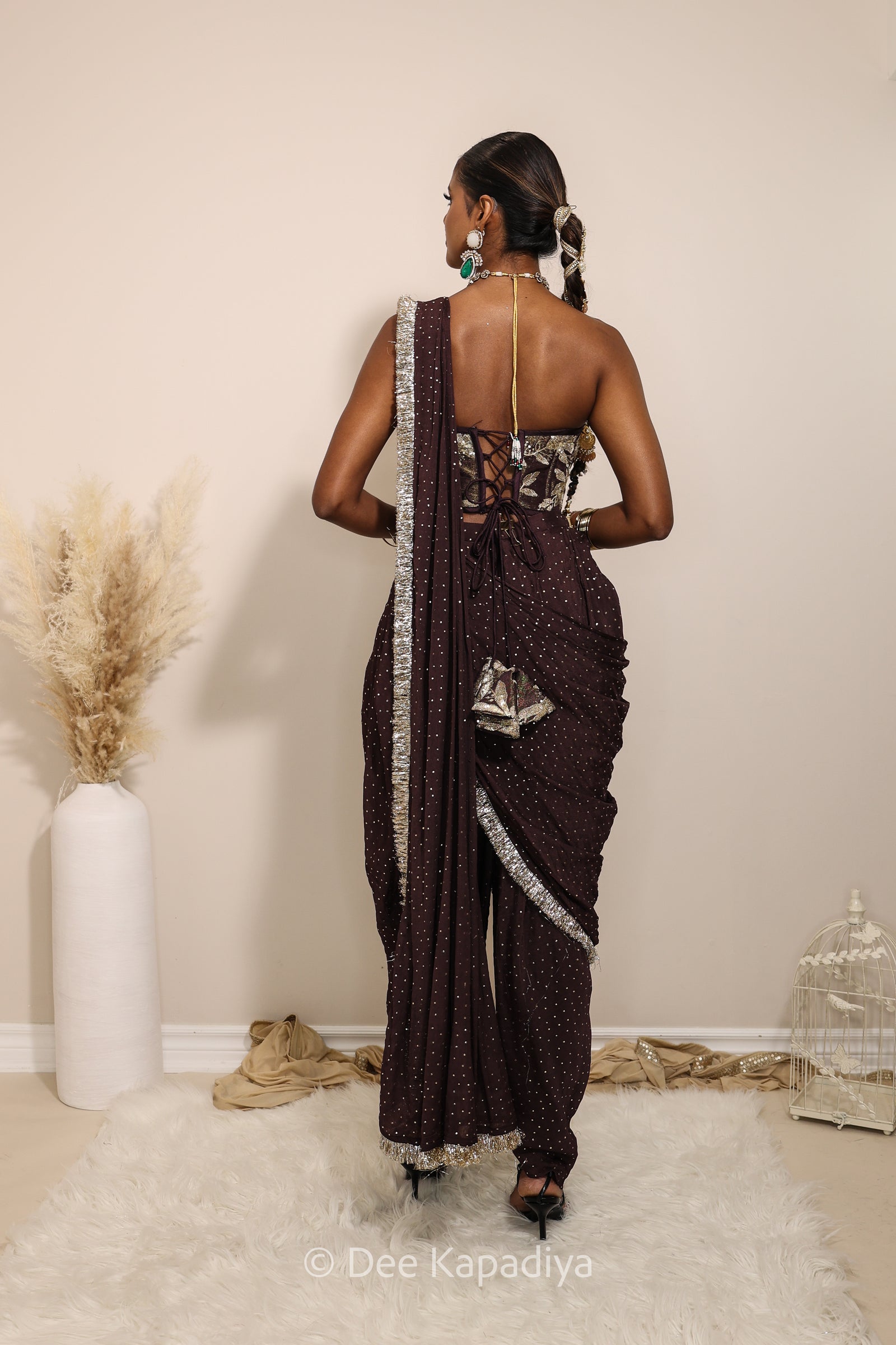 Woman in a floral brown and gold/silver patterns traditional dhoti saree outfit with jewelry, standing against a plain background. Ahilya corset top with Devi Dhoti saree front in espresso plum chocolate brown for wedding, bridal haldi, mehendi jaggo, sangeet indian wedding events 