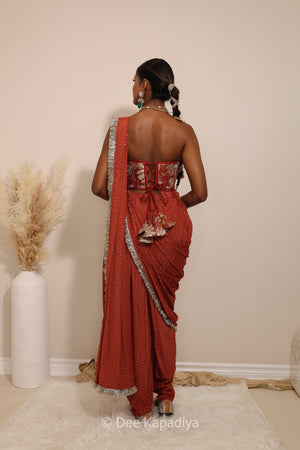 Woman in a floral red and gold/silver patterns traditional dhoti saree outfit with jewelry, standing against a plain background. Ahilya corset top with Devi Dhoti saree back in sunkissed rust red for wedding, bridal haldi, mehendi jaggo, sangeet indian wedding events 