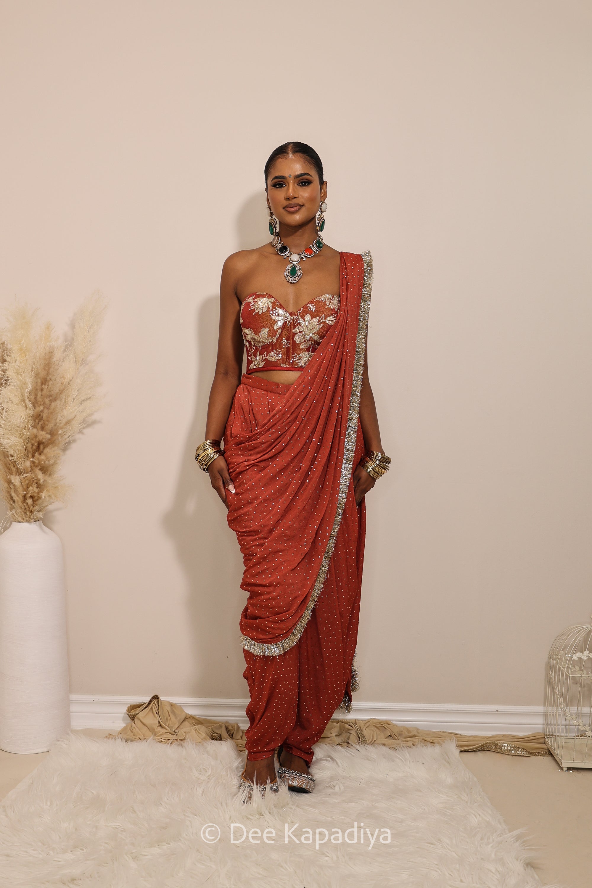 Woman in a floral red and gold/silver patterns traditional dhoti saree outfit with jewelry, standing against a plain background. Ahilya corset top with Devi Dhoti saree back in sunkissed rust red for wedding, bridal haldi, mehendi jaggo, sangeet indian wedding events 