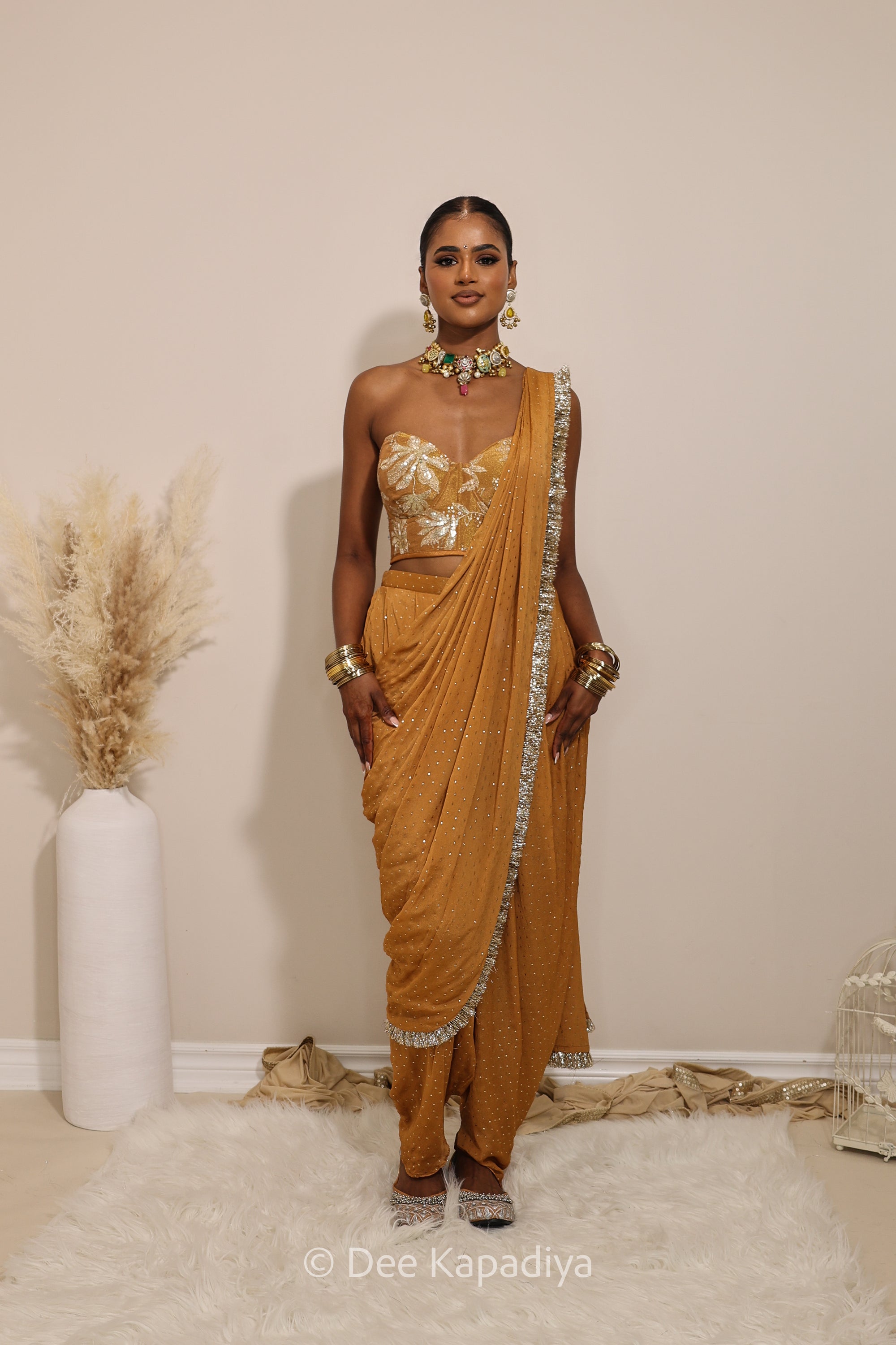 Woman in a floral yellow and gold/silver patterns traditional dhoti saree outfit with jewelry, standing against a plain background. Ahilya corset top with Devi Dhoti saree front in amber sienna mustard yellow for wedding, bridal haldi, mehendi jaggo, sangeet indian wedding events 