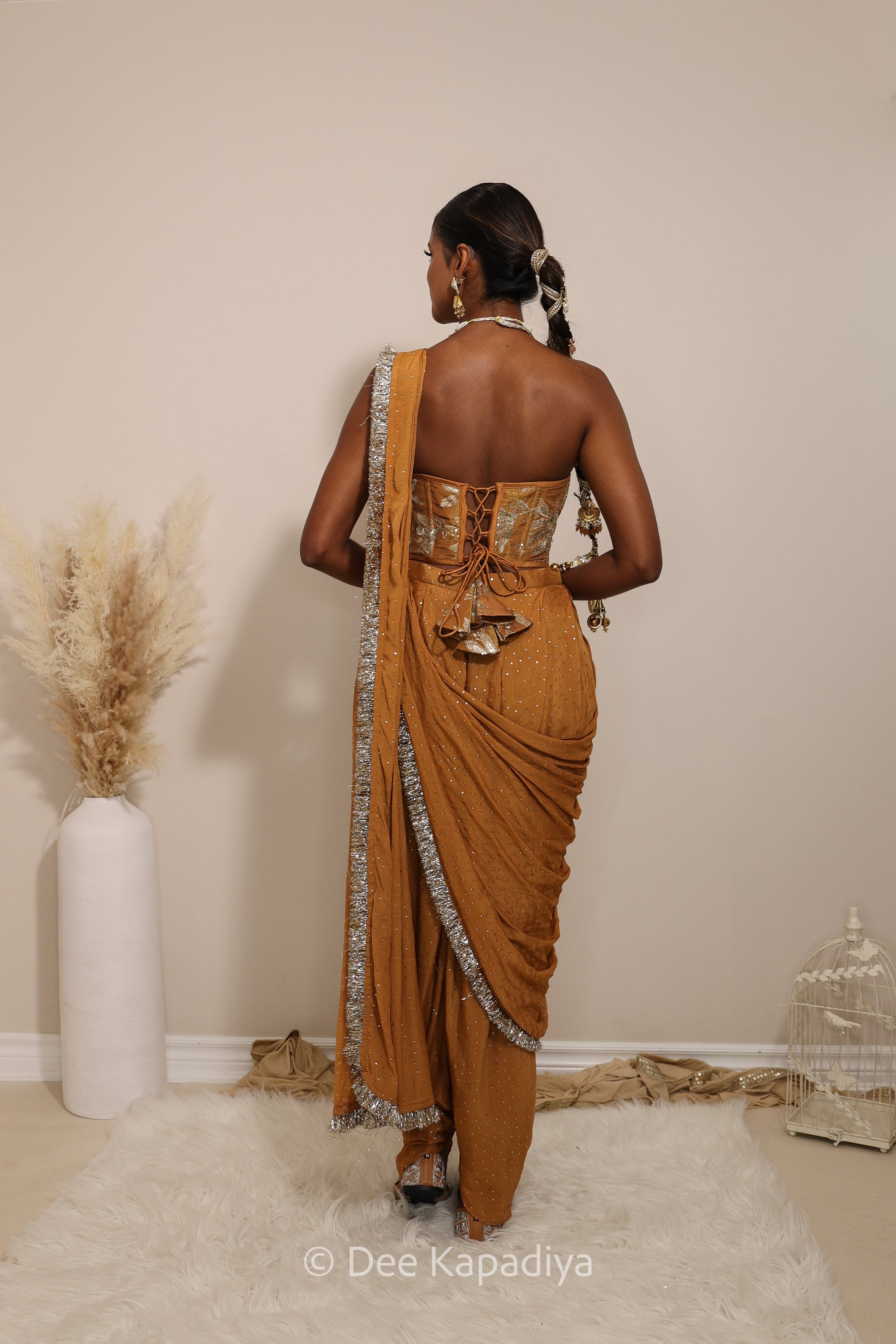 Woman in a floral yellow and gold/silver patterns traditional dhoti saree outfit with jewelry, standing against a plain background. Ahilya corset top with Devi Dhoti saree front in amber sienna mustard yellow for wedding, bridal haldi, mehendi jaggo, sangeet indian wedding events 