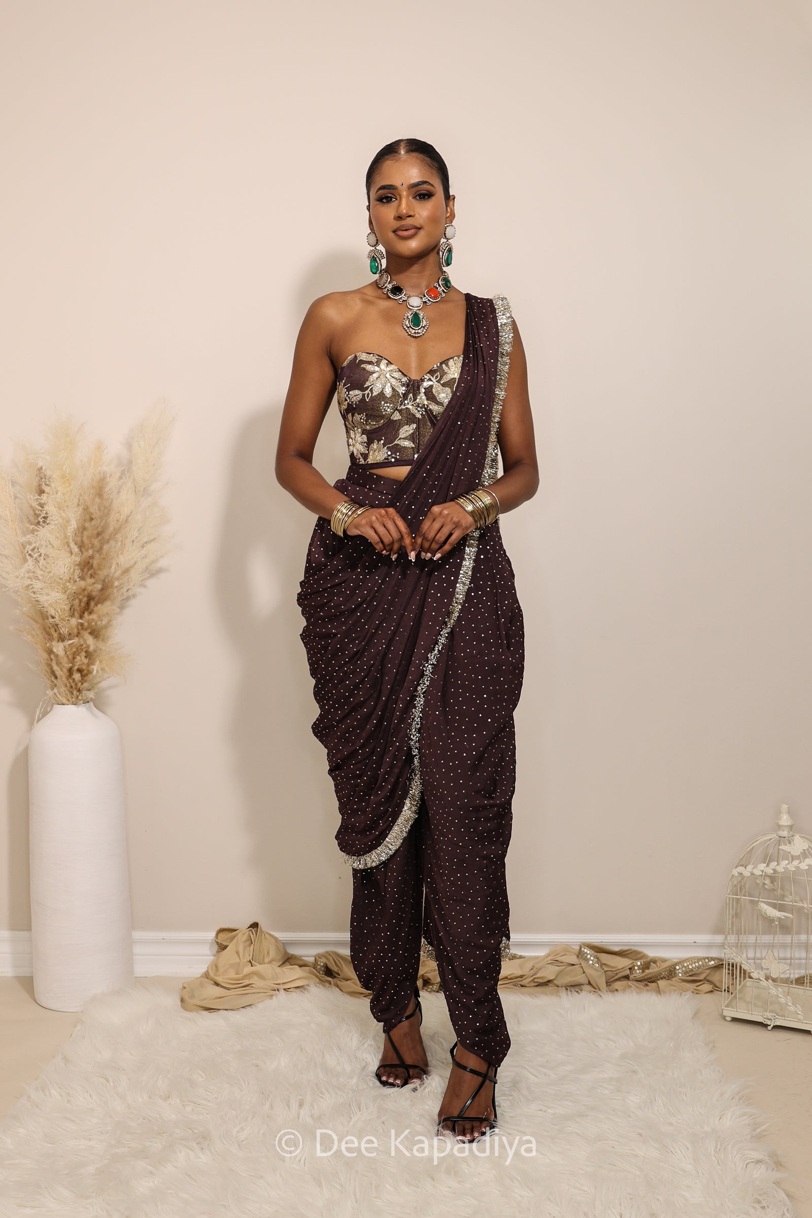 Woman in a floral brown and gold/silver patterns traditional dhoti saree outfit with jewelry, standing against a plain background. Ahilya corset top with Devi Dhoti saree front in espresso plum chocolate brown for wedding, bridal haldi, mehendi jaggo, sangeet indian wedding events 