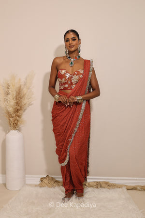 Woman in a floral red and gold/silver patterns traditional dhoti saree outfit with jewelry, standing against a plain background. Ahilya corset top with Devi Dhoti saree front in sunkissed rust red for wedding, bridal haldi, mehendi jaggo, sangeet indian wedding events 