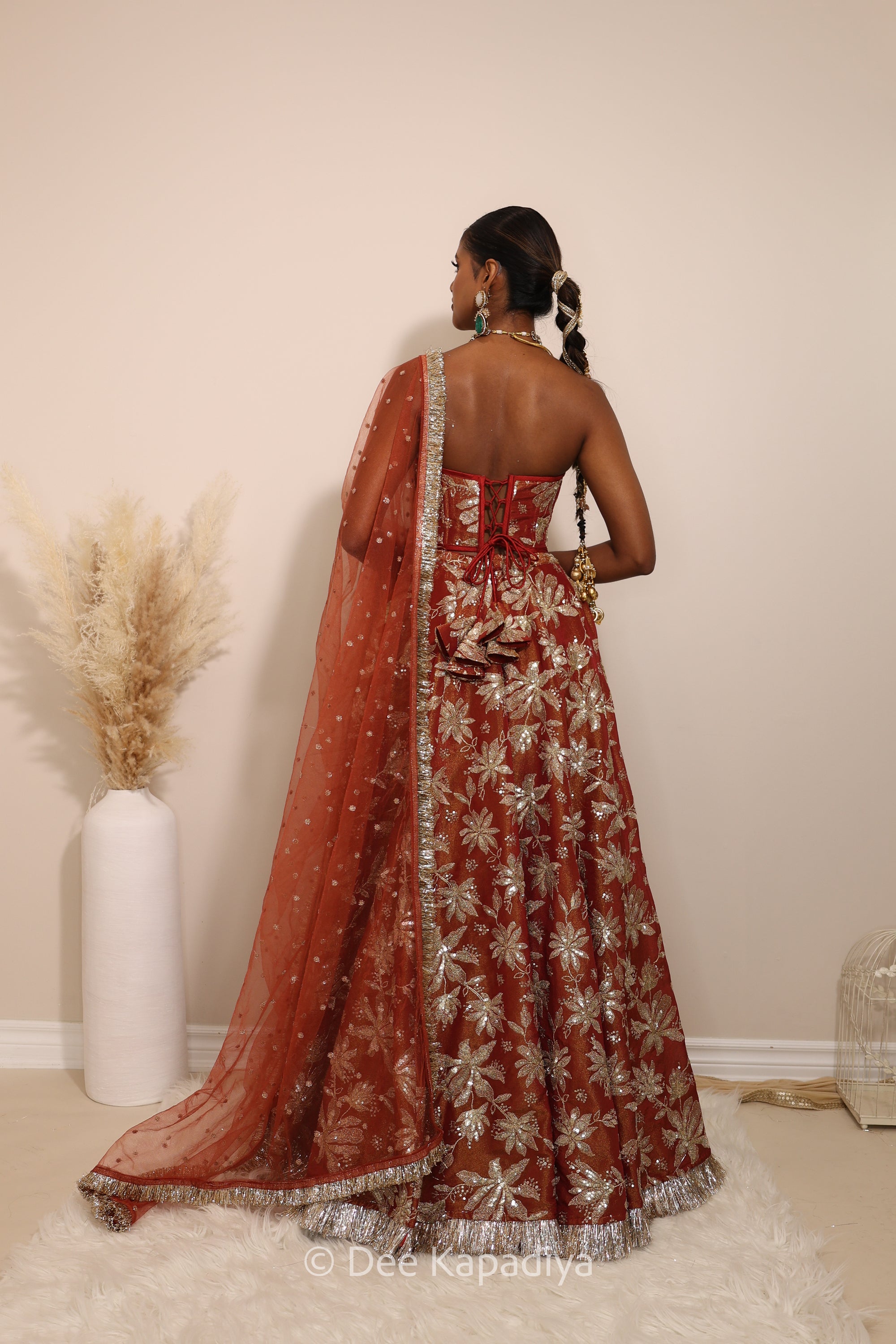Woman in a floral red classic lehenga with gold patterns with a sheer dupatta standing against a neutral background. Ahilya corset top with Jodha Lehenga back in sunkissed rust red for wedding, bridal haldi, mehendi jaggo, sangeet indian wedding events
