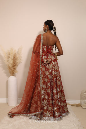 Woman in a floral red classic lehenga with gold patterns with a sheer dupatta standing against a neutral background. Ahilya corset top with Jodha Lehenga back in sunkissed rust red for wedding, bridal haldi, mehendi jaggo, sangeet indian wedding events
