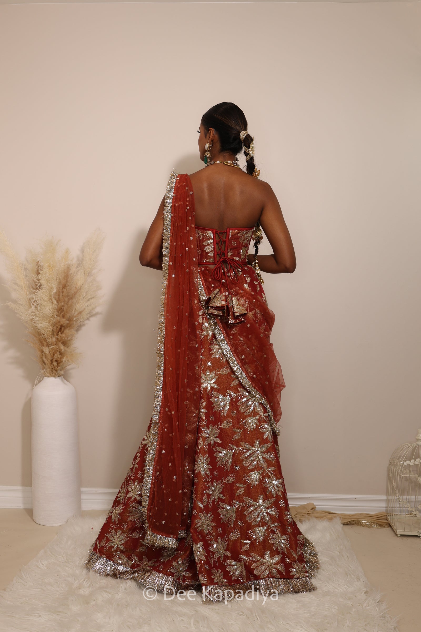 Woman in a floral red mermaid lehenga with gold patterns with a sheer dupatta standing against a neutral background. Ahilya corset top with Vaani marmaid Lehenga front in sunkissed rust red for wedding, bridal haldi, mehendi jaggo, sangeet indian wedding events