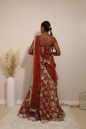 Woman in a floral brown and gold/silver patterns traditional mermaid lehenga outfit with jewelry, with a sheer dupatta standing against a plain background. Ahilya corset top with Vaani marmaid Lehenga back in sunkissed rust red for wedding, bridal haldi, mehendi jaggo, sangeet indian wedding events 