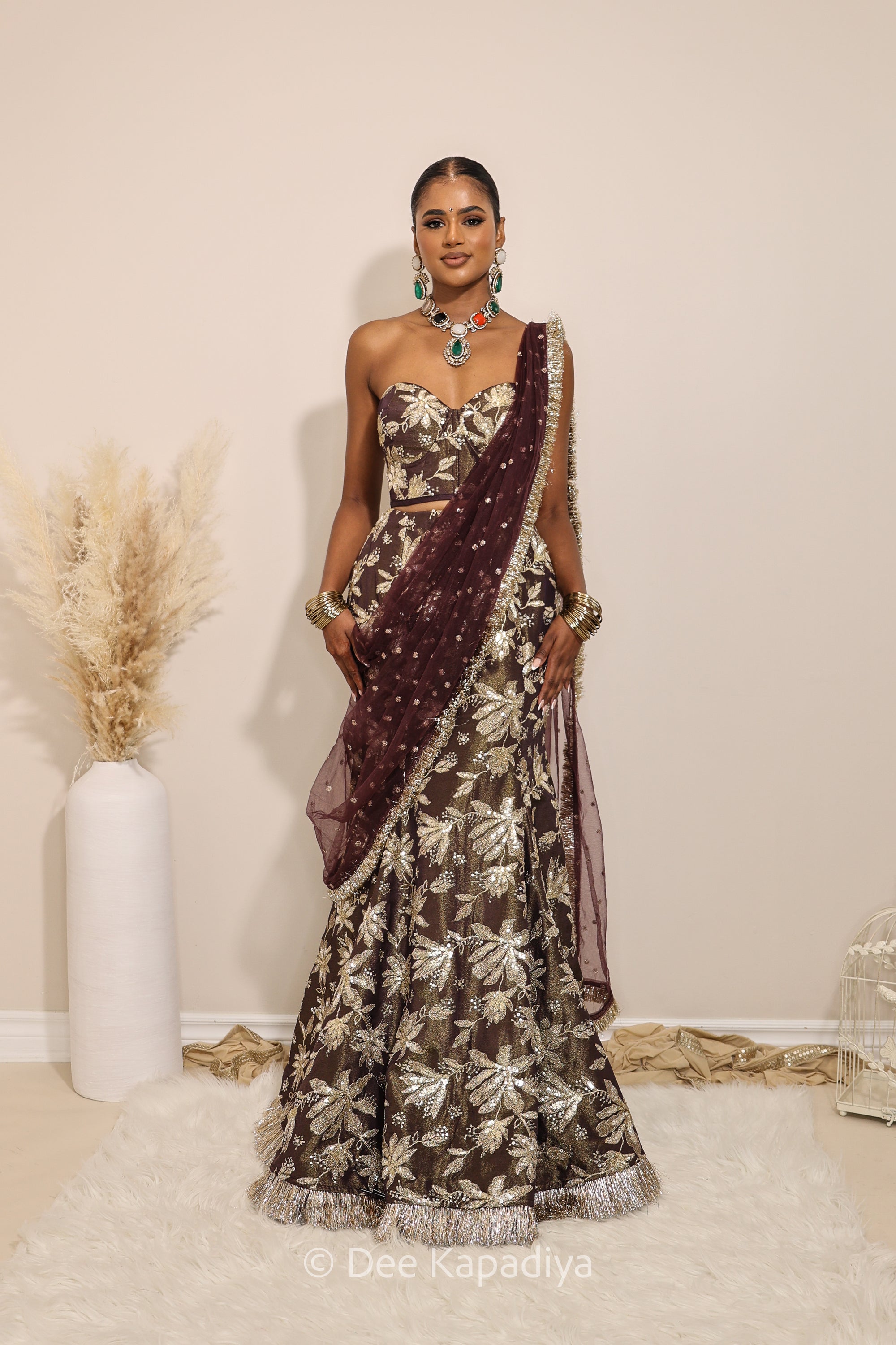 Woman in a floral brown and gold/silver patterns traditional mermaid lehenga outfit with jewelry, with a sheer dupatta standing against a plain background. Ahilya corset top with Vaani marmaid Lehenga front in espresso plum chocolate brown for wedding, bridal haldi, mehendi jaggo, sangeet indian wedding events 