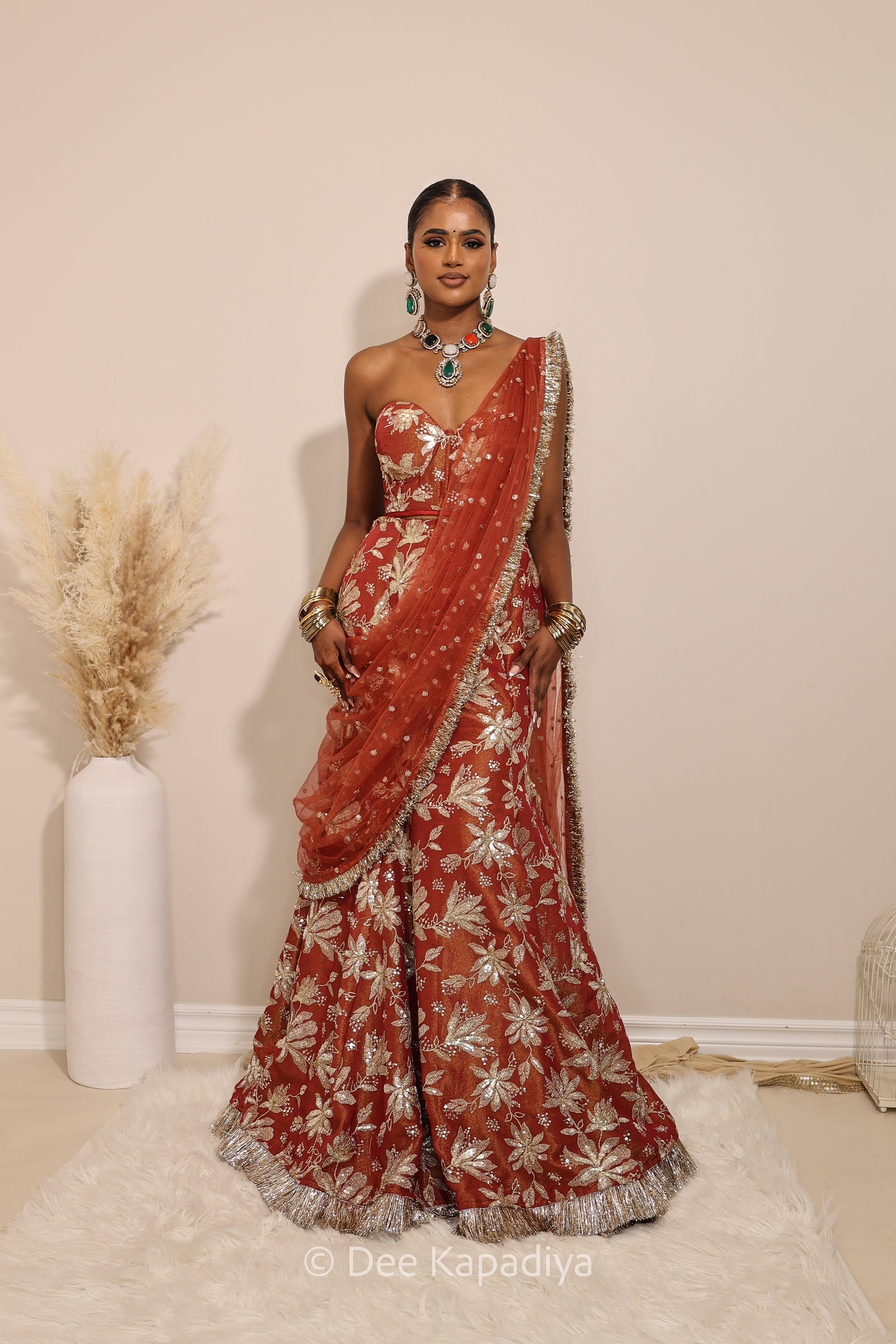 Woman in a floral red mermaid lehenga with gold patterns with a sheer dupatta standing against a neutral background. Ahilya corset top with Vaani marmaid Lehenga front in sunkissed rust red for wedding, bridal haldi, mehendi jaggo, sangeet indian wedding events