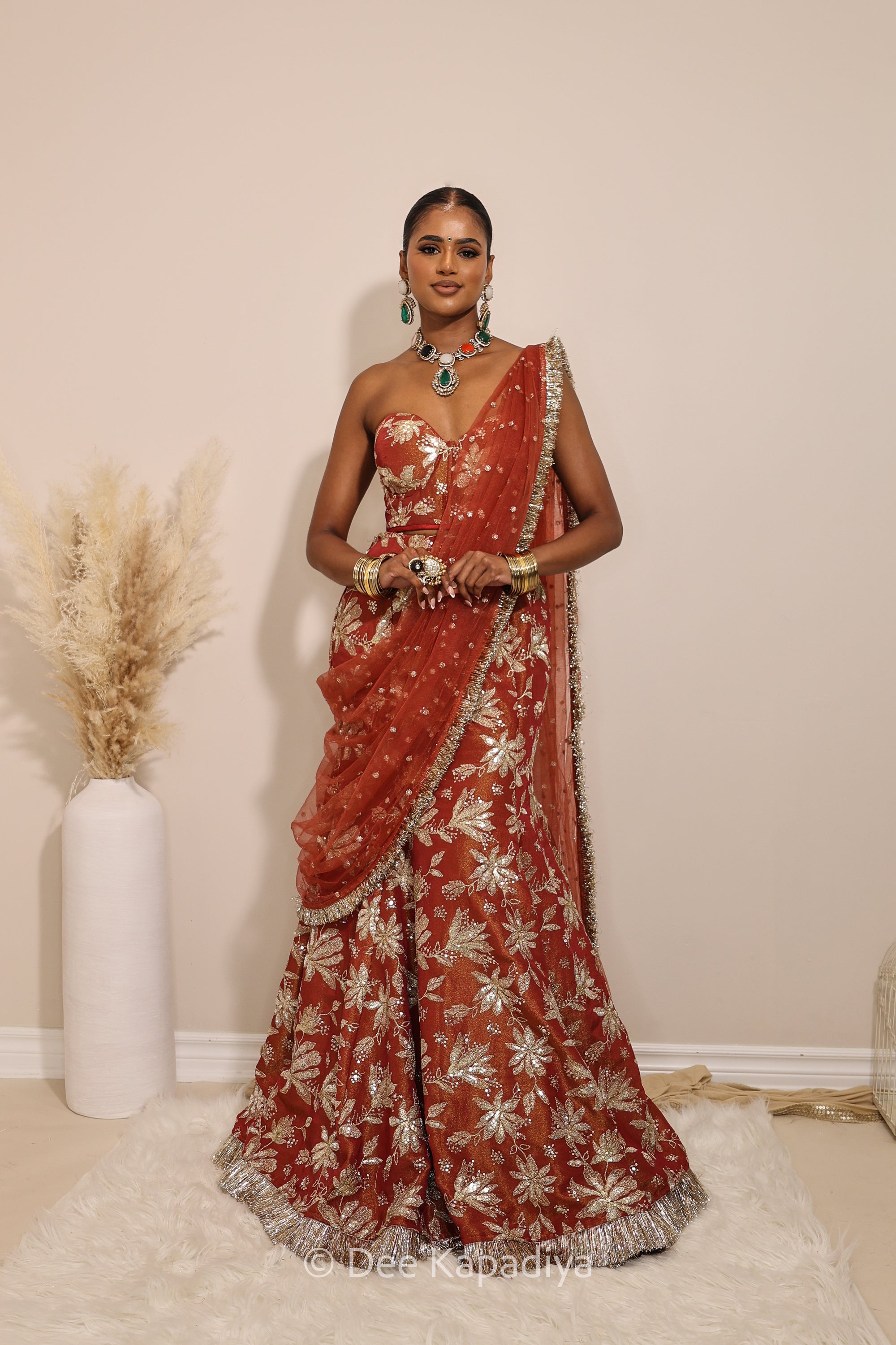 Woman in a floral brown and gold/silver patterns traditional mermaid lehenga outfit with jewelry, with a sheer dupatta standing against a plain background. Ahilya corset top with Vaani marmaid Lehenga front in sunkissed rust red for wedding, bridal haldi, mehendi jaggo, sangeet indian wedding events 