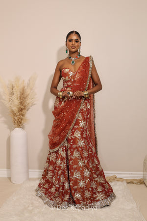 Woman in a floral brown and gold/silver patterns traditional mermaid lehenga outfit with jewelry, with a sheer dupatta standing against a plain background. Ahilya corset top with Vaani marmaid Lehenga front in sunkissed rust red for wedding, bridal haldi, mehendi jaggo, sangeet indian wedding events 