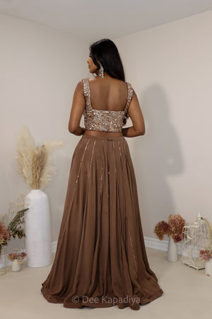 A woman stands with her back to the camera wearing a two-piece brown formal lehenga with a heavily embellished broze sequined crop top and a flowing pleated skirt. The photo is taken in a minimalist room with beige walls, decorated with pampas grass in a white vase and dried flowers.