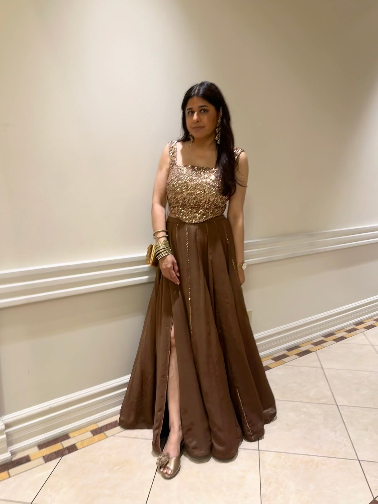A Dee Kapadiya woman wearing a floor-length brown evening lehenga with a sequined bronze corset, standing in an indoor hallway with cream walls and tiled floors.