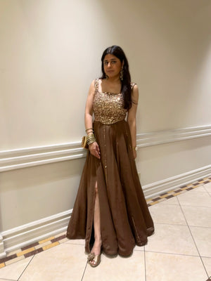 A Dee Kapadiya woman wearing a floor-length brown evening lehenga with a sequined bronze corset, standing in an indoor hallway with cream walls and tiled floors.