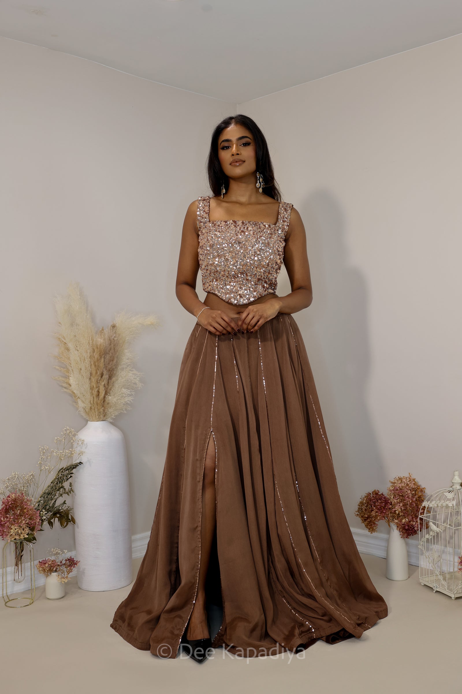 A Dee Kapadiya woman wearing a floor-length brown evening lehenga with a sequined bronze corset, standing in an indoor hallway with cream walls and tiled floors.