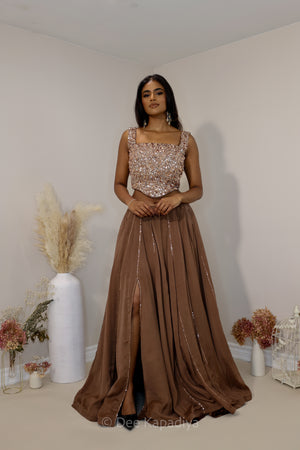 A Dee Kapadiya woman modeling a floor-length evening lehenga with a heavily embellished metallic bronze sequined corset and a flowing taupe-brown skirt/lehenga with slit, standing in a decorated indoor room