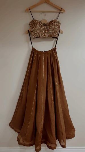 A two-piece brown formal lehenga croptop hanging on a wooden hanger against a plain off-white wall. The top is a spagatte strap sweetheart-style bodice covered in dense bronze sequins, and the panel skirt is a flowing brown chiffon thin vertical bronze stripes.