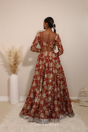Woman in a floral lehenga skirt with a sheer dupatta standing against a neutral background. Lakshmi top with Jodha Lehenga back in sunkissed rust red for wedding, bridal haldi, mehendi jaggo, sangeet indian wedding events