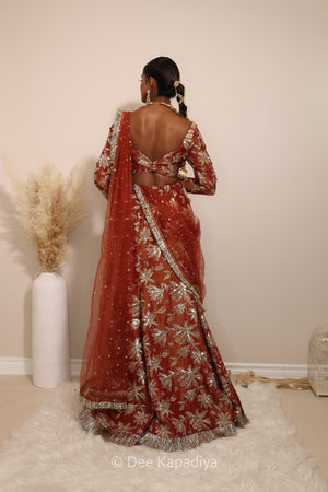 Woman in a floral red mermaid lehenga with gold patterns with a sheer dupatta standing against a neutral background. Lakshmi top with Vaani Lehenga back in sunkissed rust red for wedding, bridal haldi, mehendi jaggo, sangeet indian wedding events