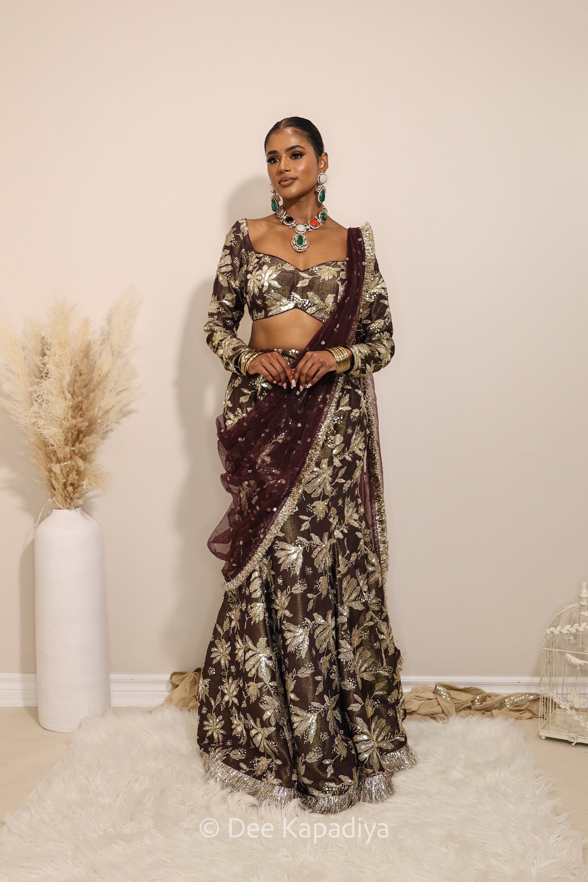 Woman in a floral brown mermaid lehenga with gold patterns with a sheer dupatta standing against a neutral background. Lakshmi top with Vaani Lehenga front in espresson plum chocolate brown for wedding, bridal haldi, mehendi jaggo, sangeet indian wedding events