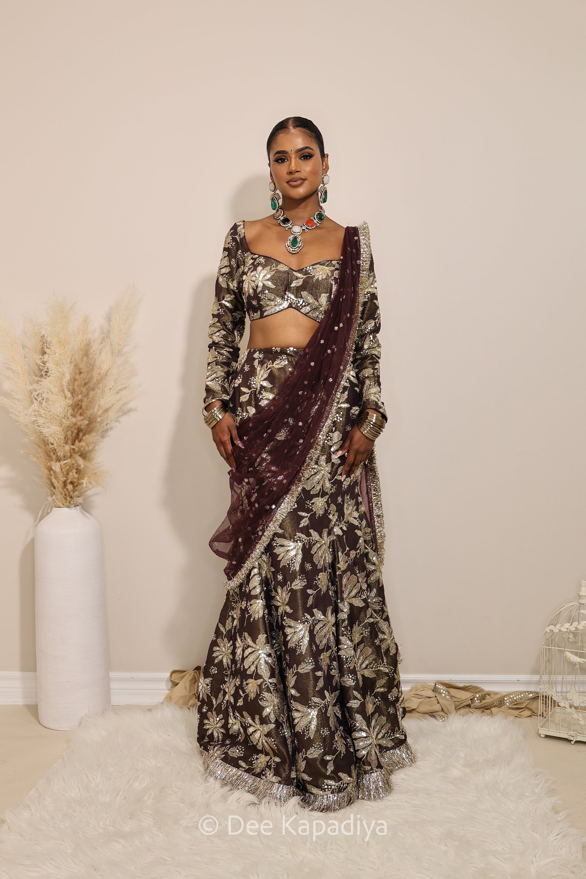 Woman in a floral brown mermaid lehenga with gold patterns with a sheer dupatta standing against a neutral background. Lakshmi top with Vaani Lehenga front in espresson plum chocolate brown for wedding, bridal haldi, mehendi jaggo, sangeet indian wedding events