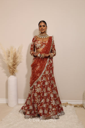 Woman in a floral red and gold traditional mermaid lehenga outfit with jewelry, with a sheer dupatta standing against a plain background. Lakshmi top with Vaani marmaid Lehenga front in sunkissed rust red for wedding, bridal haldi, mehendi jaggo, sangeet indian wedding events