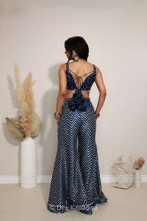 A woman stands with her back to the camera wearing a navy indigo blue and white chevron and bandhani printed patterned two-piece set with a cutout back and tie detail. The setting is a minimalist room with beige walls, a white vase with dried pampas grass, and a small decorative birdcage.