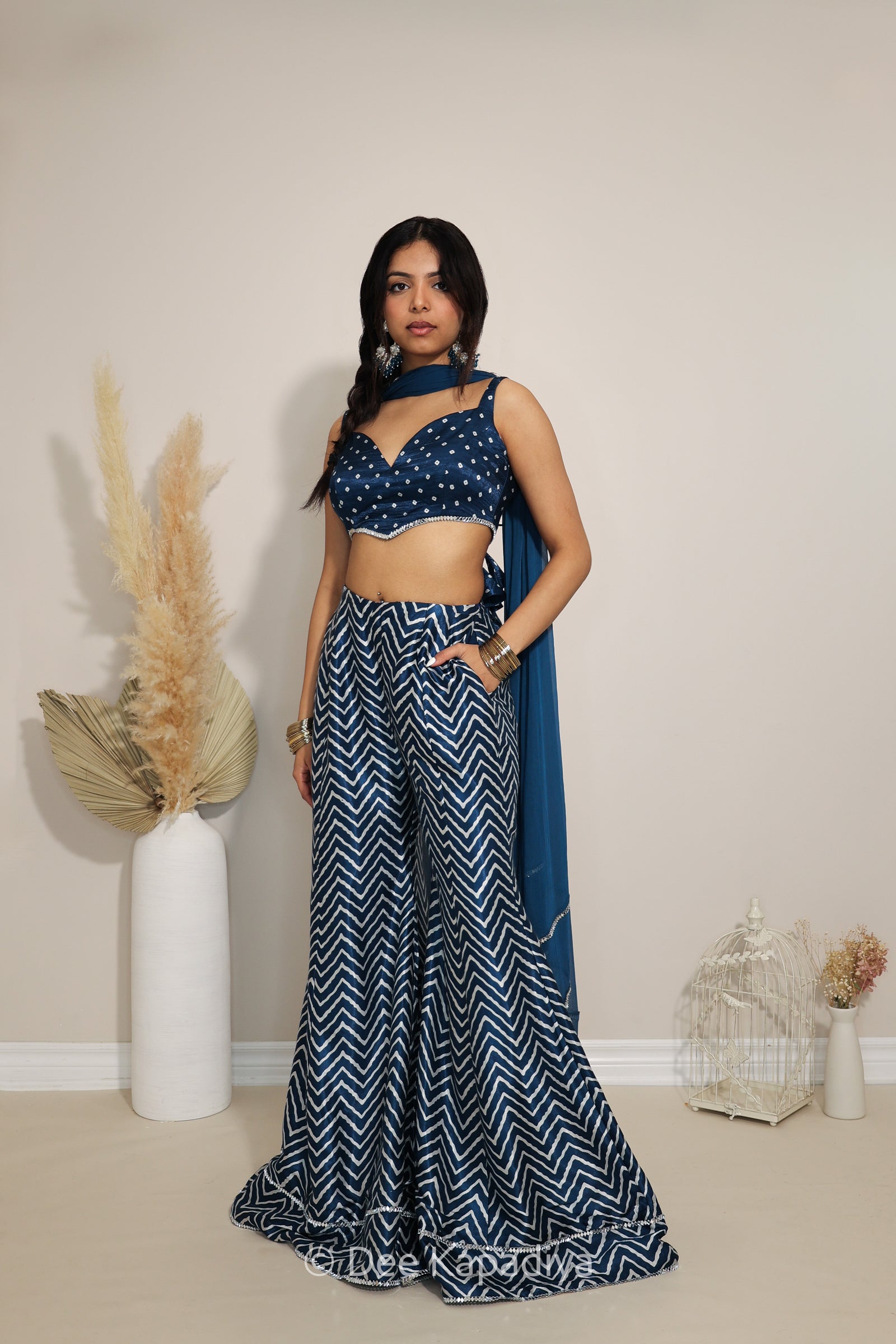A woman models a navy indigo blue and white printed bandhani and chevron traditional outfit consisting of a crop top, wide-leg pants, and a dupatta, standing in a studio decorated with pampas grass and a birdcage.