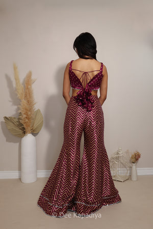 A woman viewed from behind wearing a deep burgundy flared pant with a chevron pattern and a ruffled back bandhani print detail, standing in a room with neutral beige walls and decorative pampas grass.