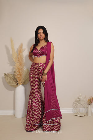 A woman models a magenta and white chevron-patterned Indian ethnic outfit, consisting of a sleeveless bandhani print crop top, flared pants, and a draped dupatta, set against a neutral beige wall with decorative pampas grass.