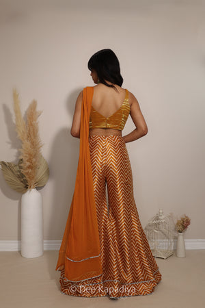 A woman stands with her back to the camera wearing a traditional Indian outfit consisting of a gold and yellow chevron-patterned skirt, a matching gold blouse, and an yellow dupatta. The photo is taken in a studio setting with decorative dried pampas grass in white vases.