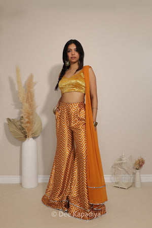 A woman modeling a traditional Indian outfit consisting of a metallic gold crop top, wide-leg trousers with a gold and yellow chevron pattern, and a solid yellow dupatta, standing in a studio with decorative dried grass and a small birdcage.