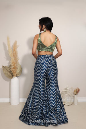 A woman shown from the back wearing a two-piece outfit consisting of a green camouflage crop top and navy blue wide-leg pants with a white chevron pattern, standing in a studio decorated with dried pampas grass.