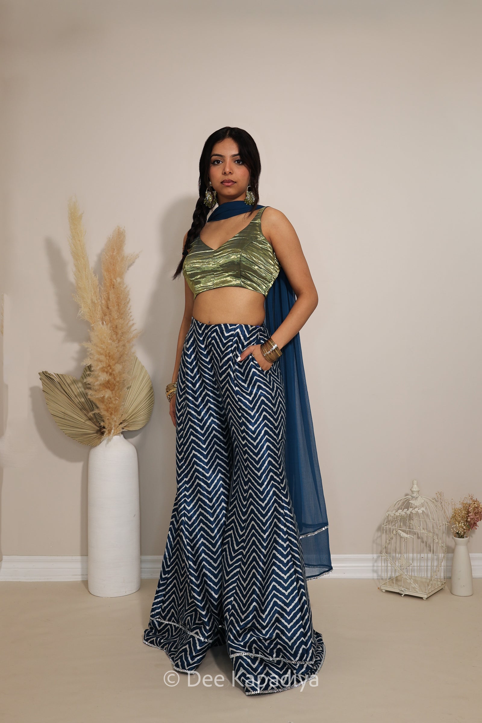 A woman models a two-piece ethnic outfit consisting of a metallic olive green crop top and navy blue wide-leg pants with a white chevron pattern, paired with a matching navy blue dupatta. She is standing in a studio setting with decorative dried pampas grass in a white vase on the left.