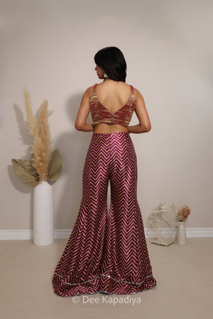 A woman stands with her back to the camera wearing a sleeveless maroon and gold chevron-patterned two-piece outfit with a deep V-back top and matching flared pants. The photo is taken in a studio with neutral beige walls and floor, decorated with pampas grass in a white vase and a small white birdcage.