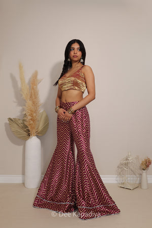 A woman modeling a two-piece ethnic wear set consisting of a metallic gold crop top and maroon and gold chevron patterned flared pants, standing in front of a beige wall with decorative pampas grass in a white vase.