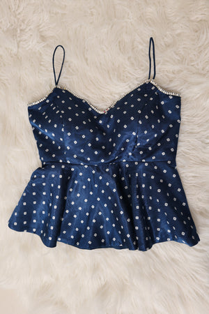 A indigo peplum top with white bandhani print and thin spaghetti straps, laid flat on a white faux fur background.
