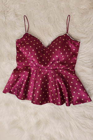 A burgundy peplum top with white bandhani print and thin spaghetti straps, laid flat on a white faux fur background.