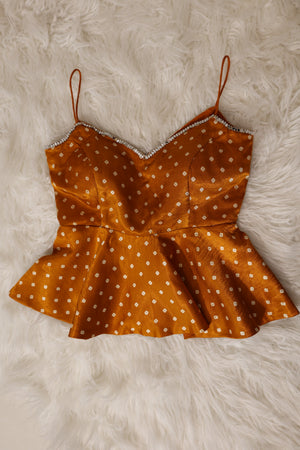 An orange haldi yellow peplum top with white bandhani print and thin spaghetti straps, laid flat on a white faux fur background.
