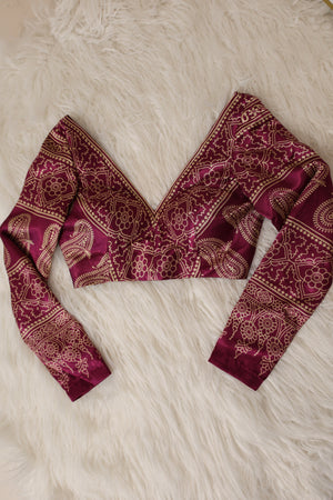A long-sleeve V-neck blouse with an intricate maroon and gold paisley ajrakh pattern, laid flat on a white faux fur background.
