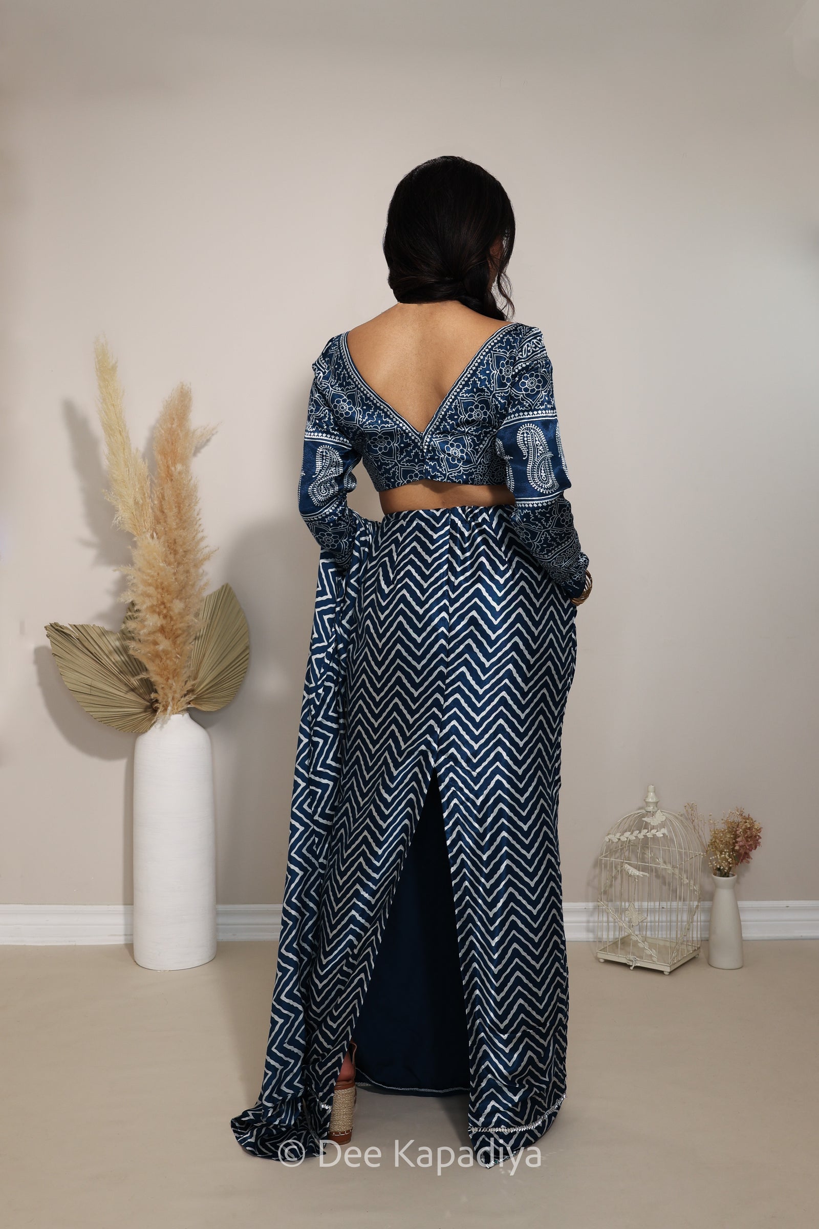 A woman modeling a navy blue and white geometric patterned two-piece outfit with a long sleeve ajrakh print blouse and pre-draped saree in chevron print, standing in a neutral indoor setting with decorative dried grass and a birdcage.