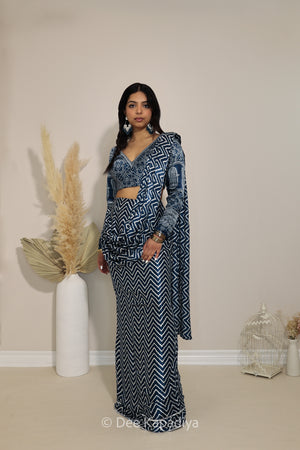 A woman models a matching two-piece outfit featuring a long-sleeve ajrakh print crop top and a pre-draped chevron printed saree, both decorated with a navy blue and white geometric chevron pattern. She is standing in a room with a beige wall, next to a white vase with dried pampas grass.