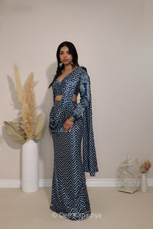 A woman modeling a navy blue and white geometric patterned two-piece outfit with a long sleeve ajrakh print blouse and pre-draped saree in chevron print, standing in a neutral indoor setting with decorative dried grass and a birdcage.