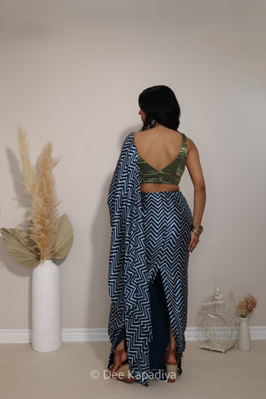 A woman stands with her back to the camera wearing a indigo blue and white metallic tissue v neck croptop with pre-draped chevron print pattern saree with a green velvet crop blouse. She is standing in a room with a beige wall, decorated with a tall white vase containing dried pampas grass on the left and a small white wire birdcage with dried flowers on the right.