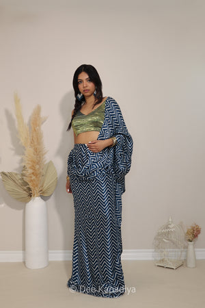 A woman modeling a indigo blue and white patterned two-piece outfit with a metallic olive green crop top metallic tissue v neck croptop with pre-draped chevron print pattern saree, standing in a studio with decorative dried grass in a white vase and a small birdcage on a beige wall.