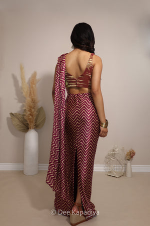 A woman is shown from behind wearing a maroon and white chevron-patterned metallic tissue v neck croptop with pre-draped chevron print pattern saree, standing in a studio setting with decorative dried grass in a vase.