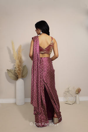 A woman stands with her back to the camera wearing a sleeveless maroon and whitemetallic tissue v neck croptop with pre-draped chevron print pattern saree with a cutout back and a matching blouse. The photo is taken in a neutral indoor setting with decorative dried plants in white vases.