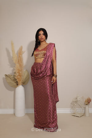 A woman modeling a maroon and white metallic tissue v neck croptop with pre-draped chevron print pattern saree  saree with a matching gold embroidered blouse, standing in a studio with decorative pampas grass and a birdcage.