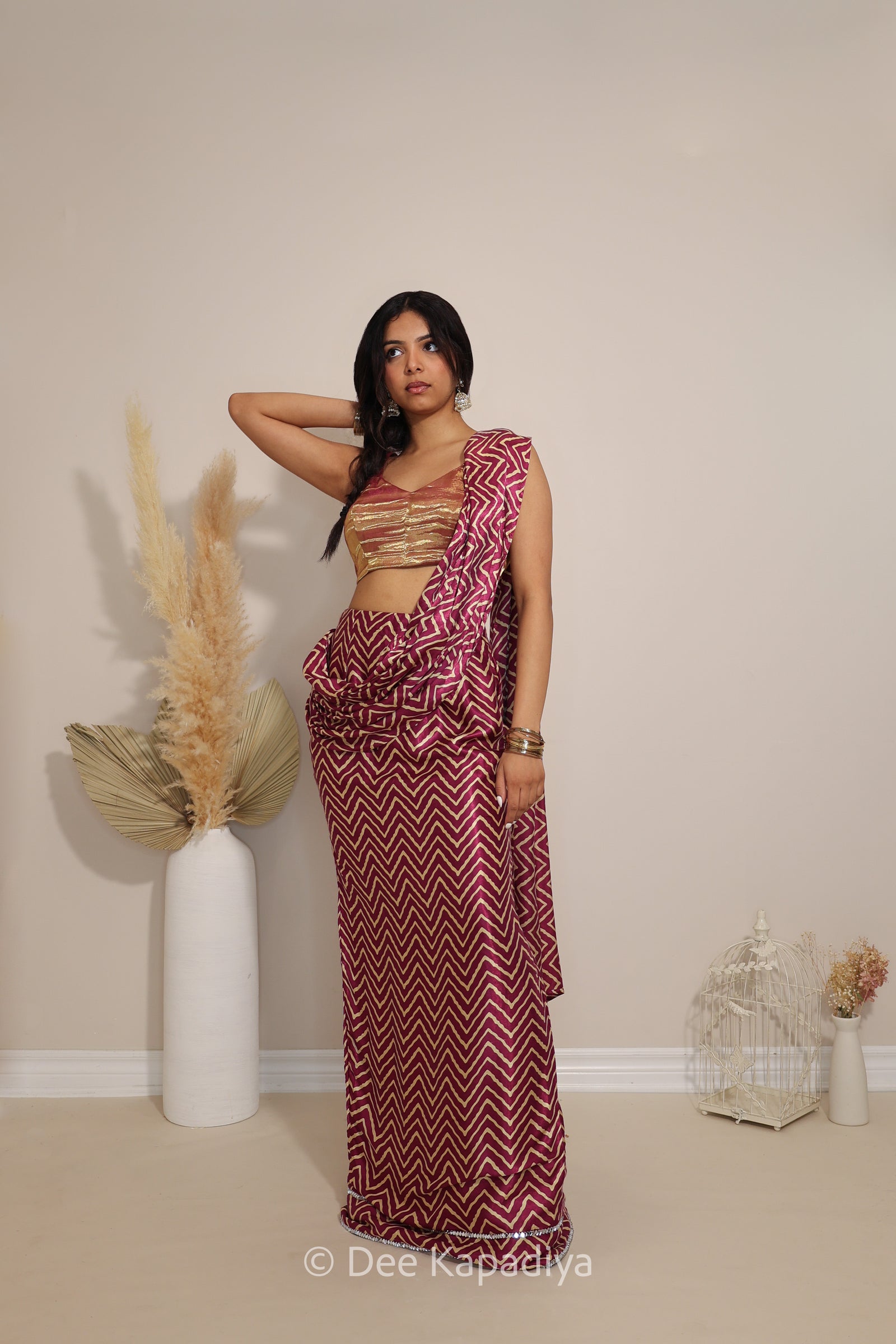 A woman models a sleeveless, metallic tissue v neck croptop with pre-draped chevron print pattern saree featuring a bold magenta and white chevron pattern, standing in a minimalist room with decorative pampas grass in a white vase.