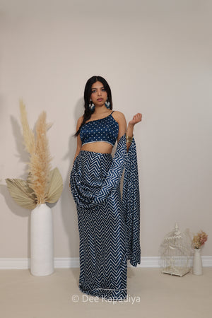 A woman modeling a navy blue and white chevron-patterned two-piece outfit consisting of a sleeveless bandhani printed 1 shoulder crop top and a draped skirt or pre-draped saree, standing in a minimalist room with decorative dried grass in a white vase and a small white birdcage.